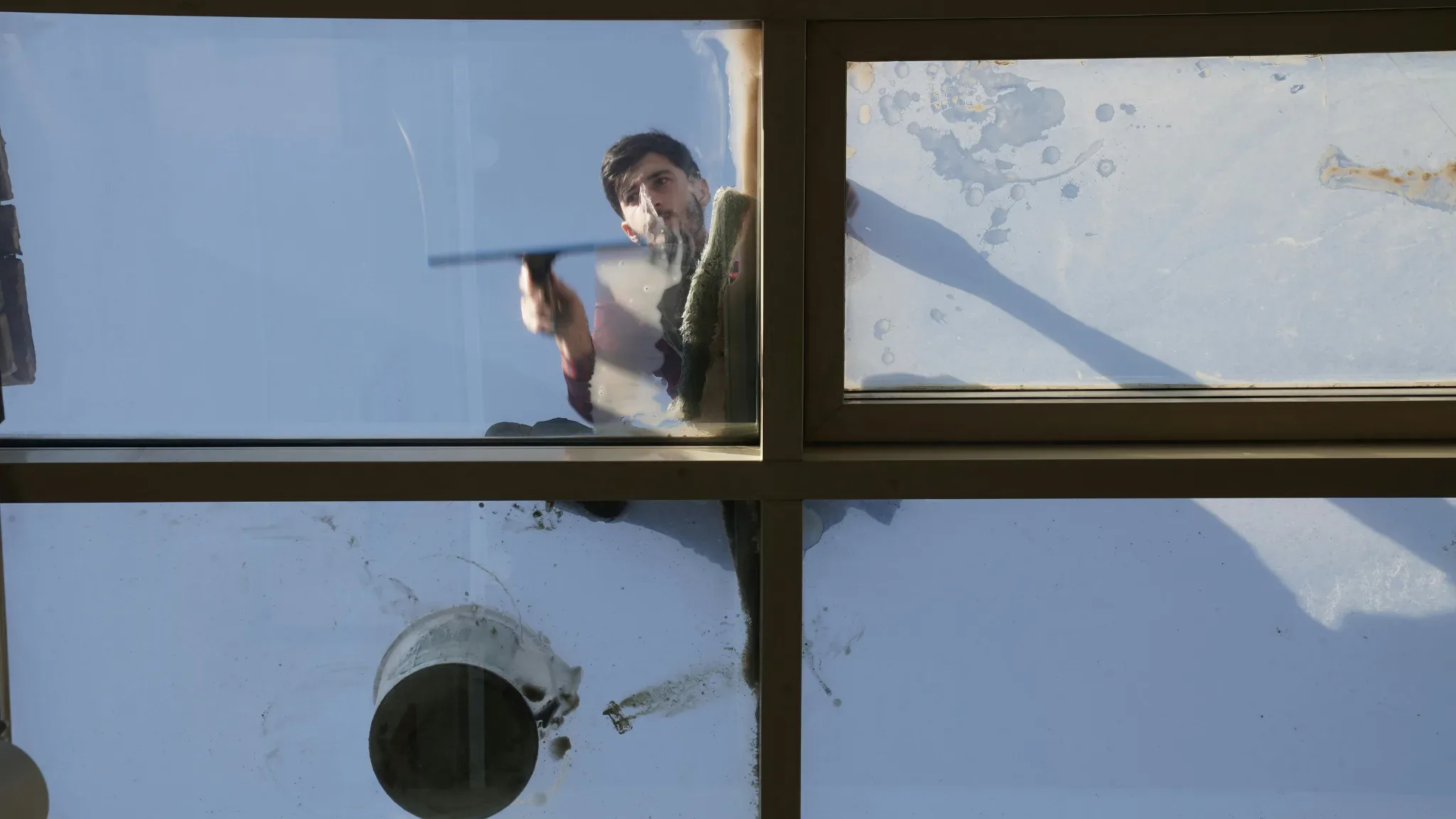 Viewed from outside, a man is seen through a large window grid, holding a small squeegee and wiping the glass on the left pan